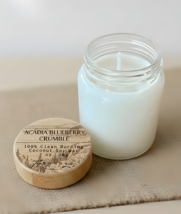 A white soy candle in a clear glass jar sits next to its engraved wooden lid on a beige textured fabric surface. The lid is printed with the scent name 'Acadia Blueberry Crumble' and the brand 'Bonded Wick'.